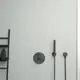 Ein schwarzer Wandthermostat von Ideal Standard mit einem Duschkopf und einem Schlauch, an einer hellen Wand montiert, neben einem schmalen Regal.